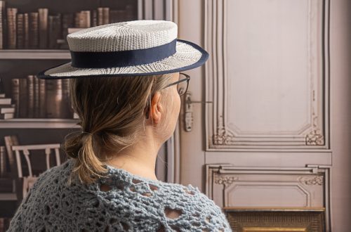 Tässä kuvassa näkyy henkilö takaapäin. Hänellä on päässään valkoinen, sinisellä nauhalla koristeltu olkihattu. Henkilöllä on yllään vaaleansininen virkattu paita tai huivi, ja hänellä on silmälasit. Taustalla näkyy kirjahylly täynnä kirjoja ja koristeellinen puinen ovi.