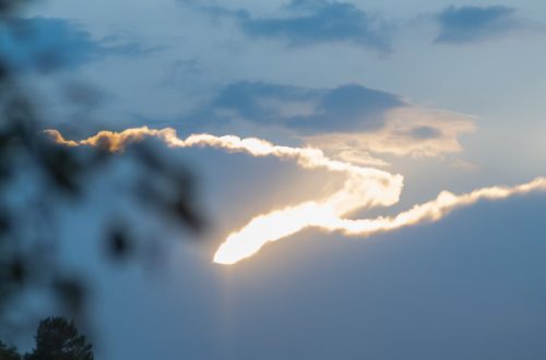 Taivaanranta auringonlaskun aikaan, jossa aurinko on osittain pilvien peitossa. Taivas hehkuu sinisen, oranssin ja keltaisen sävyissä, ja etualalla näkyy epätarkka puun tai kasvin osa.