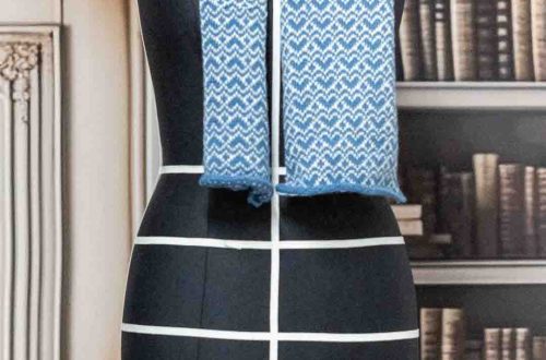 Kuvassa on musta mallinukke, jonka yllä on sinivalkoinen villakaulahuivi. Kaulahuivin kuvio on siksak-muotoinen ja se on ripustettu mallinukkeen näyttämään kaulahuivin pituutta ja kuviota.