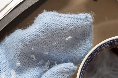 Kuva esittää asetelmaa, jossa on vaaleansininen neulottu lapanen, kuppi kahvia, kamera objektiiveineen ja avoin kirja.