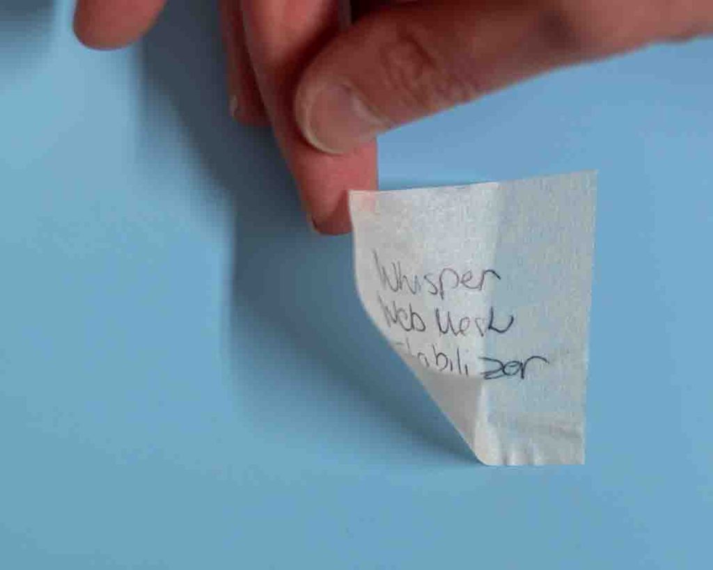 Lähikuva valkoisesta, kevyestä ja hieman läpikuultavasta kangasmaisesta palasta, joka näyttää olevan tukimateriaalia. Palassa on mustalla käsin kirjoitettu teksti 'Whisper Web Mesh Stabilizer'. Palasta pitää kiinni ihmisen sormet, ja tausta on tasainen vaaleansininen.