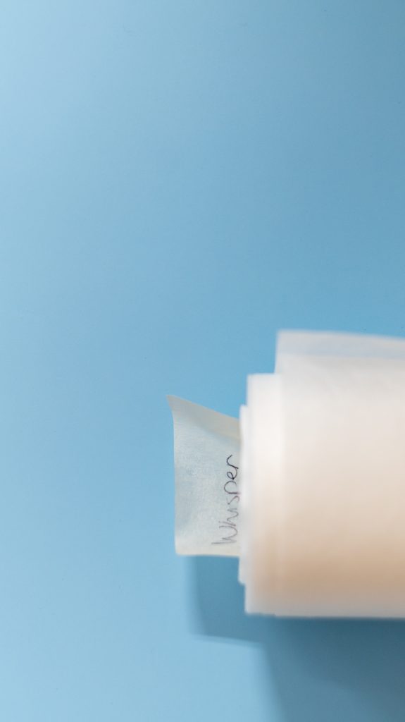 Lähikuva tukimateriaalirullasta, joka on osittain avattuna sinisellä taustalla. Rullan sisältä ulos rullautuu vaalea, läpikuultava materiaali, jonka reunaan on kiinnitetty pieni teipin pala. Teipin palassa on mustalla käsin kirjoitettu sana 'Whisper'. Kuva havainnollistaa rullan ja siihen kiinnitetyn tiedon