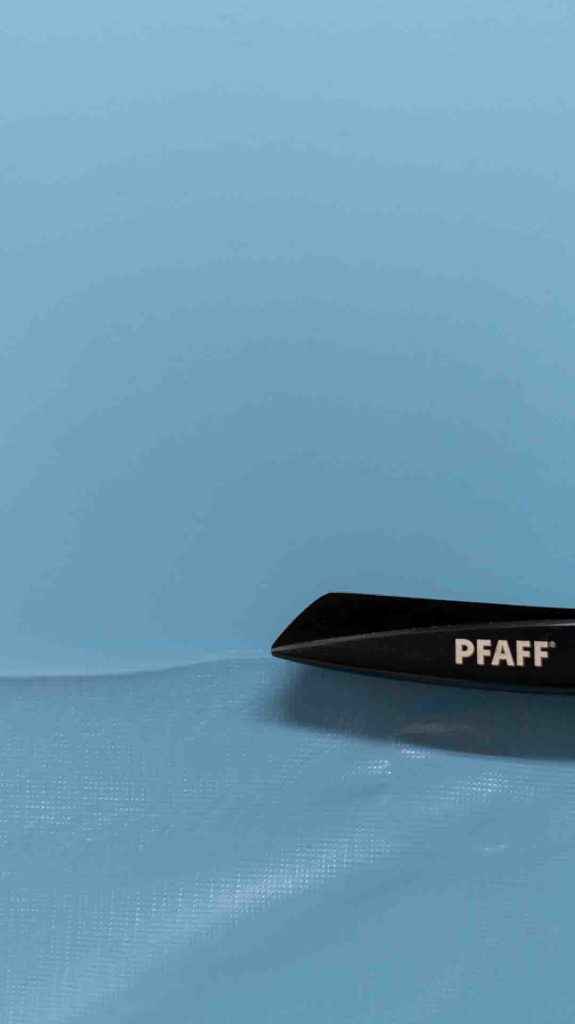 Lähikuva kirkkaan sinisellä taustalla olevasta läpinäkyvästä, hieman ryppyisestä tai aaltoilevasta kalvomaisesta materiaalista. Materiaalilla on hienojakoinen ruudukkomainen kuvio tai teksturoitu pinta. Kuvan oikeassa alakulmassa näkyy mustien saksien kärki, jossa on PFAFF-logo, leikkaamassa tai koskettamassa materiaalia.