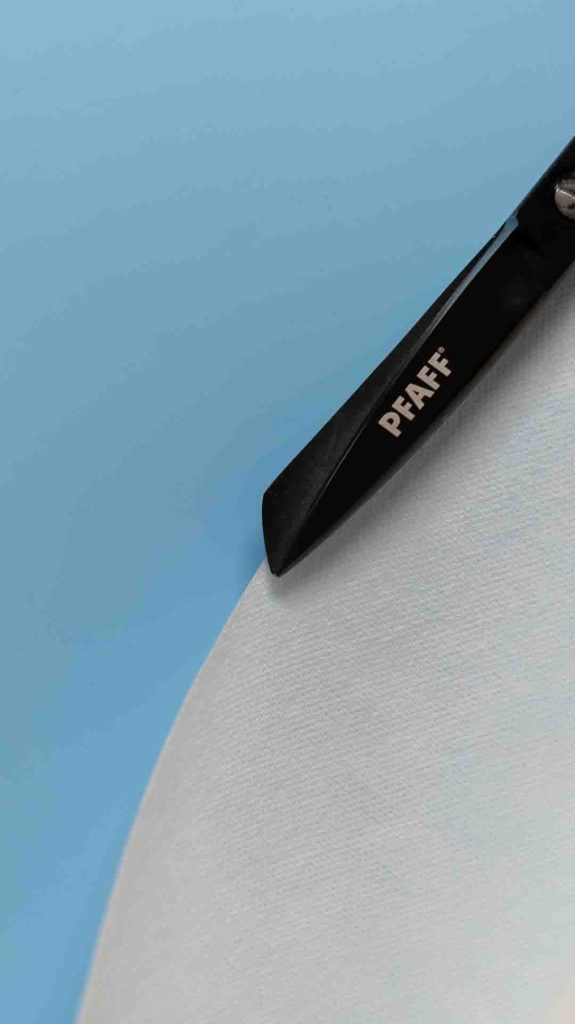 Lähikuva, jossa mustat, PFAFF-logolla varustetut sakset ovat koskettamassa tai leikkaamassa vaaleaa, paperimaista materiaalia, joka on rullalla tai kaarevasti taustalla. Materiaali näyttää olevan repäistävää tai tarrapohjaista tukimateriaalia. Tausta on tasainen vaaleansininen.