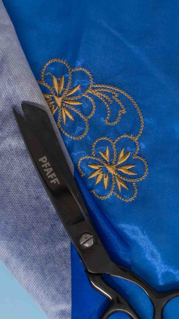 Lähikuva sinisestä satiinikankaasta, johon on brodeerattu kaksi kultaista, koristeltua kukka-aihetta. Kuvan vasemmassa reunassa näkyy osa vaaleanharmaasta leikattavasta tukimateriaalista. Kuvan keskellä on mustat ompelusaksit, joiden terä osoittaa kohti kangasta ja tukimateriaalin reunaa. Kuvassa korostuu kontrastit ja tyylikäs kirjonnan jälki.