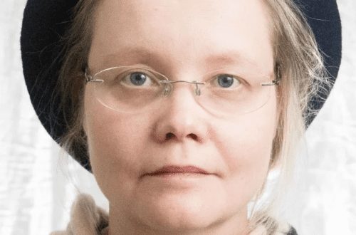Muotokuva henkilöstä, jolla on vaaleat hiukset, siniharmaat silmät ja silmälasit. Hänellä on tummansininen baskeri ja beigen, mustan ja tummanharmaan sävyinen ruudullinen huivi. Valokuva on otettu pehmeällä studiovalaistuksella, joka korostaa kasvojen piirteiden harmonista sulautumista.
