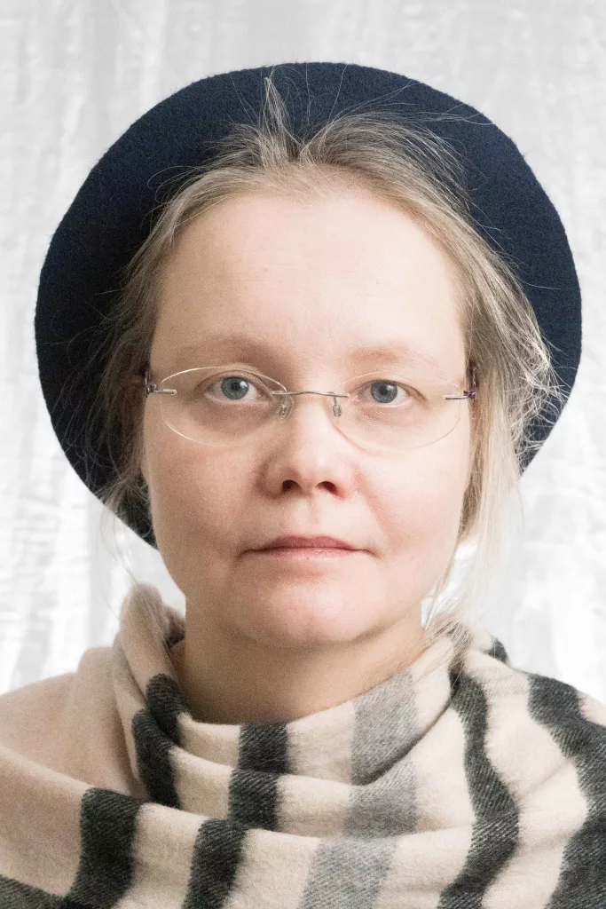 Muotokuva henkilöstä, jolla on vaaleat hiukset, siniharmaat silmät ja silmälasit. Hänellä on tummansininen baskeri ja beigen, mustan ja tummanharmaan sävyinen ruudullinen huivi. Valokuva on otettu pehmeällä studiovalaistuksella, joka korostaa kasvojen piirteiden harmonista sulautumista.