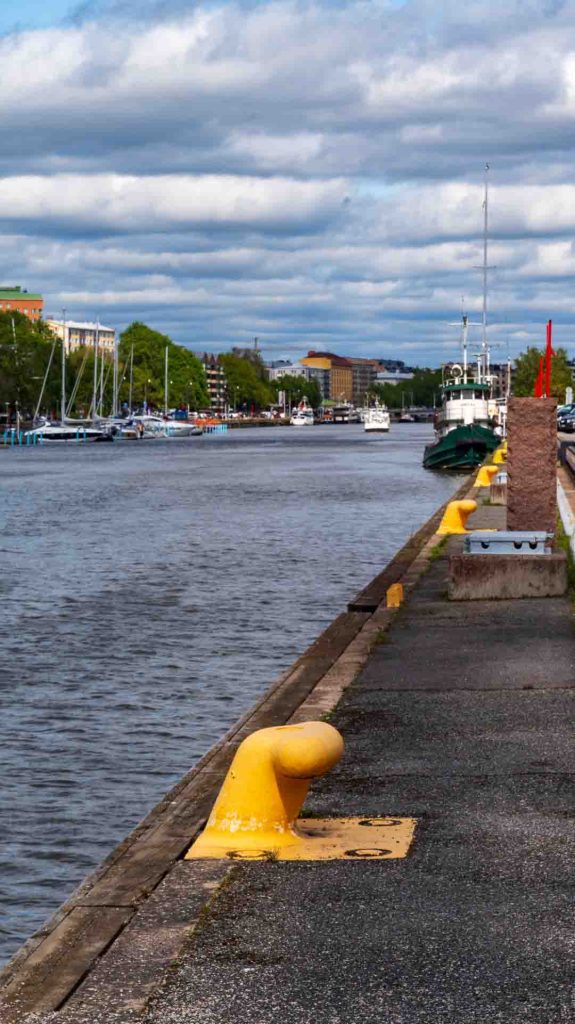 Aurajoki Turussa tyynenä kesäpäivänä, keltainen laiturin pollari etualalla ja kaupungin siluetti taustalla.