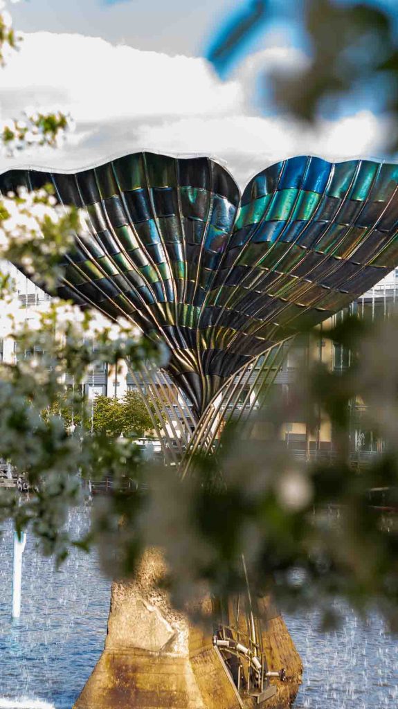 Achim Kühnin "Harmonia"-suihkulähde Turun Aurajoessa kuvattuna kukkivien oksien läpi, taustalla moderni rakennus ja pilvinen taivas.