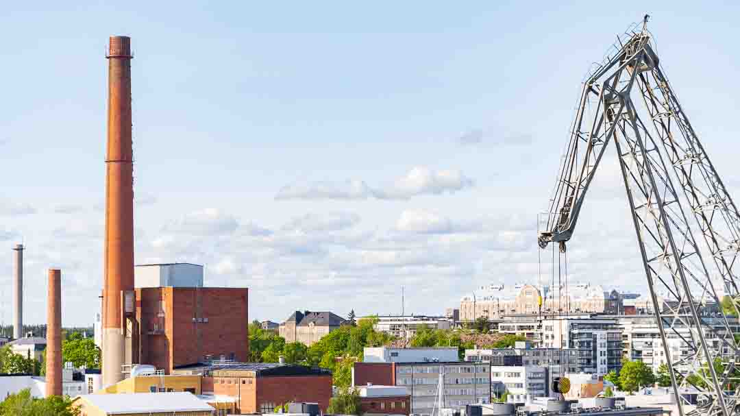 Turun kaupunkikuva, jossa näkyy korkea tiilipiippu, teollisuusrakennuksia ja suuri nosturi sinistä taivasta vasten.