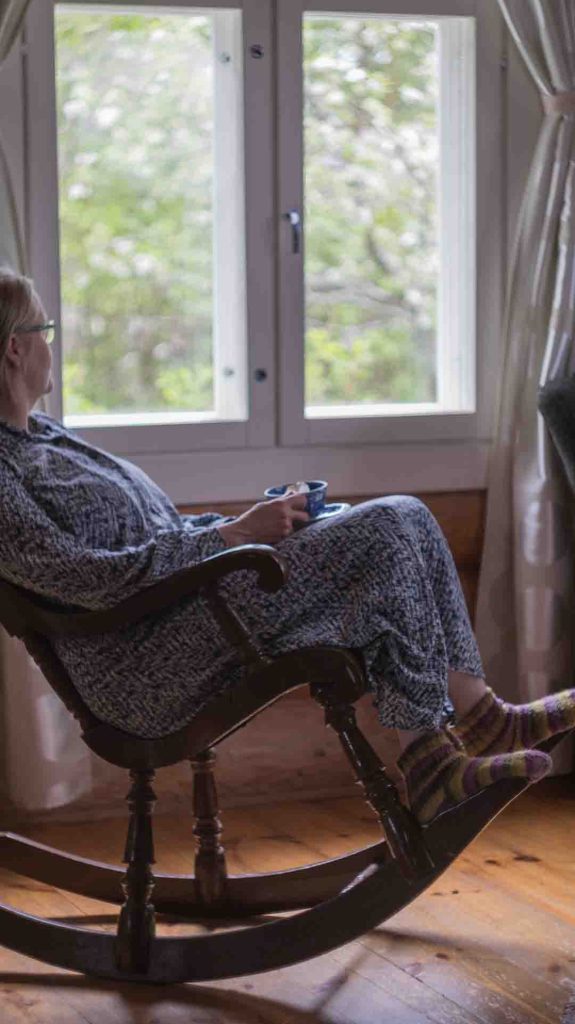 Nainen istuu puisessa keinutuolissa mökin ikkunan edessä, juo kahvia ja katsoo ulos kesäiseen maisemaan. Jaloissa on raidalliset villasukat.