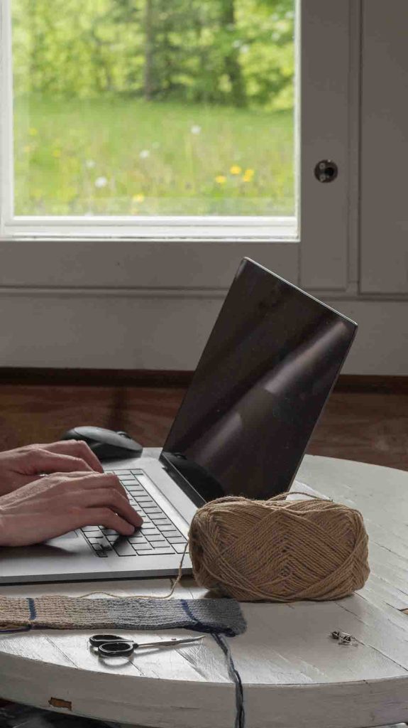 Lähikuva käsistä näppäilemässä kannettavan tietokoneen näppäimistöä mökin valkoisella pöydällä. Työpöydällä on myös lankakerä ja ompelusakset, ja taustalla näkyy vihreää luontoa ikkunan läpi.