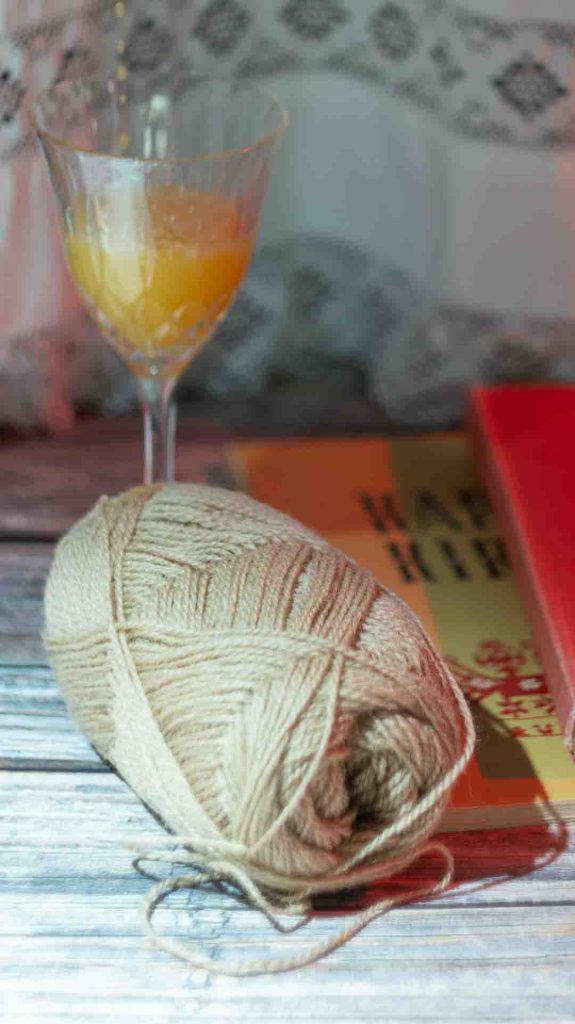  Lähikuva beigenvärisestä lankakerästä puupöydällä. Sen vieressä on oranssikantinen kirja ja lasi, jossa on oranssia juomaa. Kuvassa on pehmeä valaistus ja kodikas tunnelma.