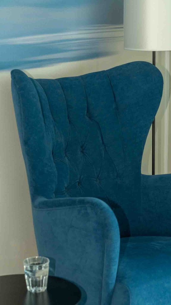 Syvän sininen, samettinen nojatuoli Imatran Valtionhotellin tai muun rauhallisen tilan nurkkauksessa. Kuva symboloi rentoutumista ja inspiraatiota.