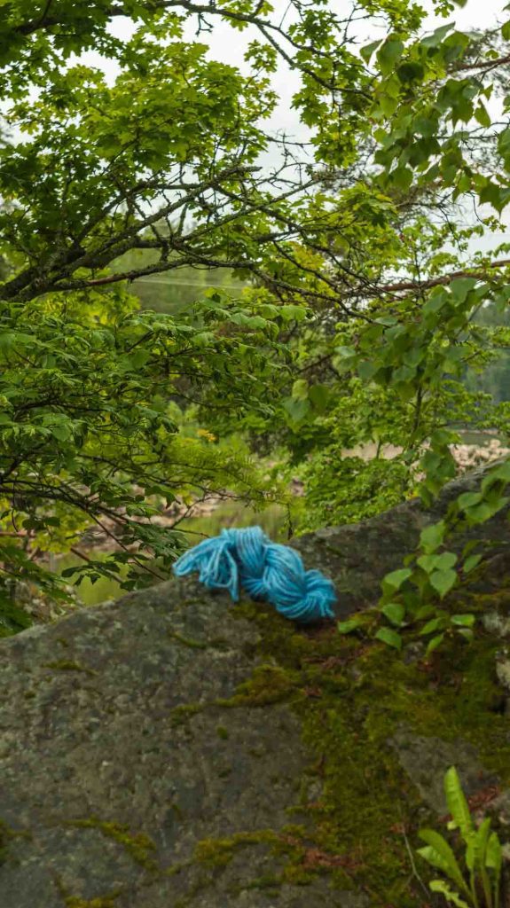 Kirkkaansininen lankakerä sammalpeitteisellä kivellä Imatran luonnossa, taustalla heijastava vesistö ja vihreää puustoa. Kuva edustaa luonnon inspiraatiota käsityöhön.