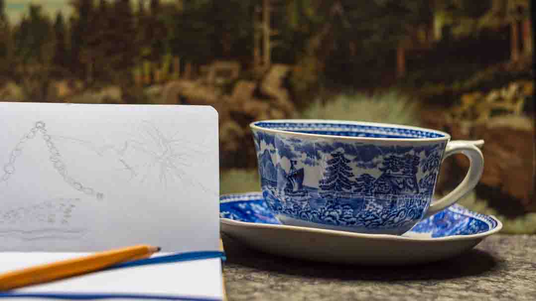 Rentouttava hetki ja luonnoskirja Imatralla. Sinivalkoinen kahvikuppi ja lyijykynällä tehty konekirjontaluonnos maisemataustalla. Kuva symboloi luovaa inspiraatiota ja mielenrauhaa käsityöläiselle.