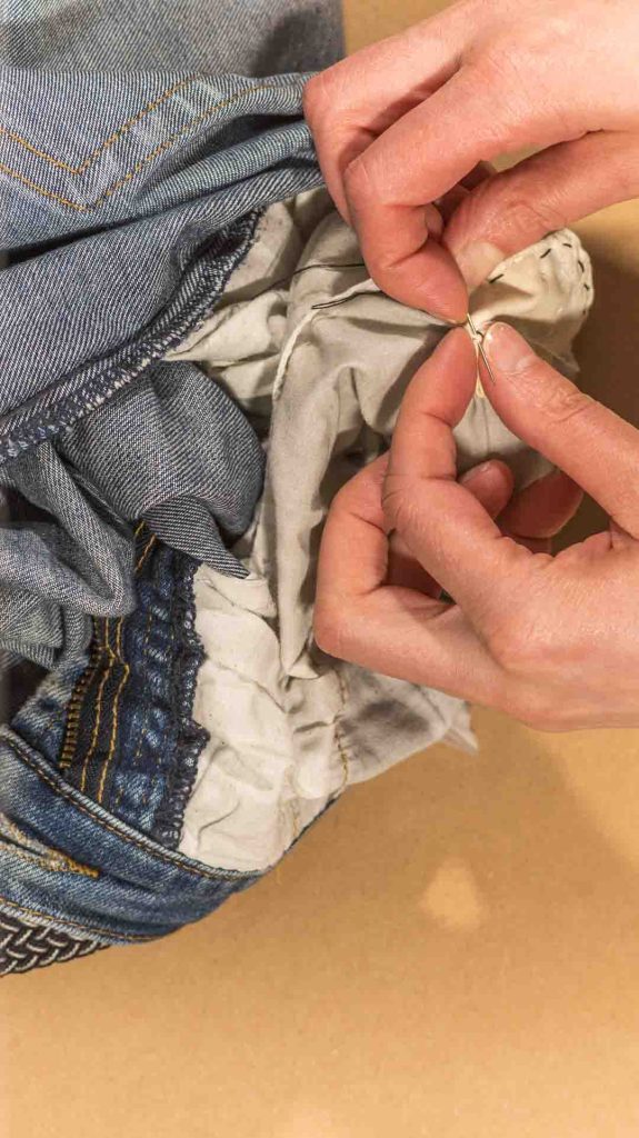 Lähikuva käsistä ompelemassa farkkua neulalla ja langalla