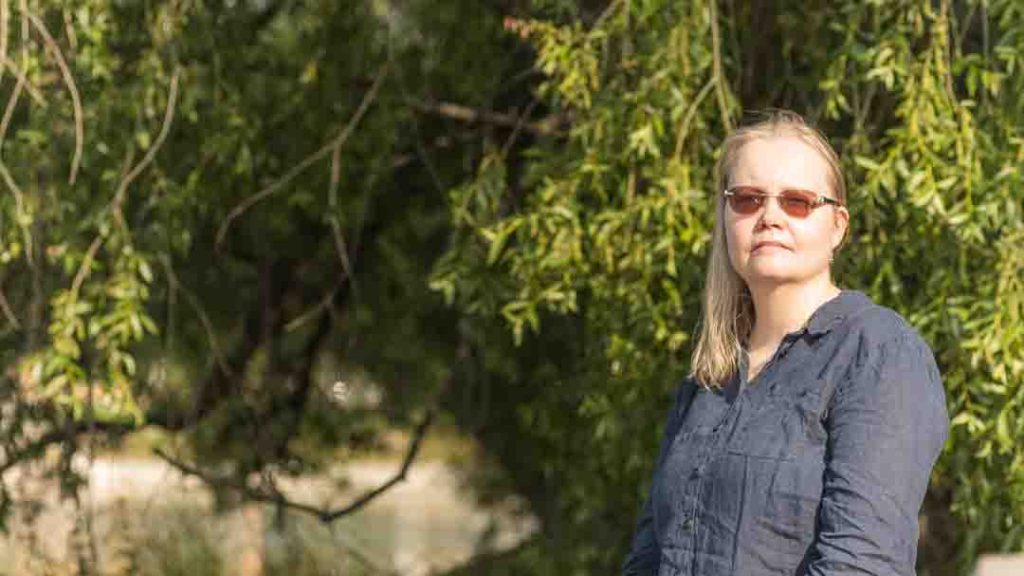 Henkilö istuu rauhallisesti Pielisjoen rannalla, katselee vettä, kuvasta huokuu pysähtymisen ja luonnon kauneuden tunnelma.