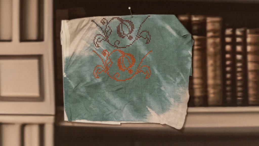 Lähikuva kirjonnan työvaiheesta, jossa tummalla langalla on tehty osa kuviosta ja kirkkaan oranssilla langalla on luonnosteltu toinen osa. Kangas on turkoosin ja valkoisen sävyinen.