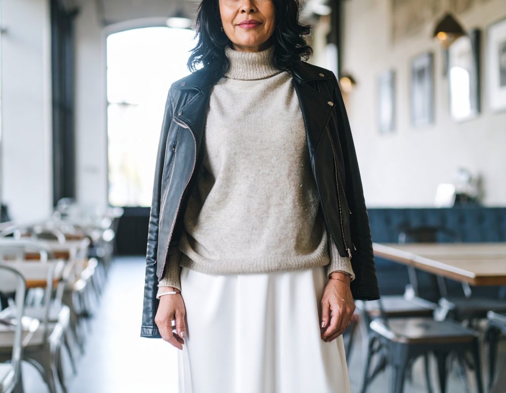 Lähikuva suomalaisesta, harmaahiuksisesta naisesta, joka esittelee taitavaa kerrospukeutumista. Hänellä on yllään paksu harmaa kaulusneule ja musta nahkatakki.