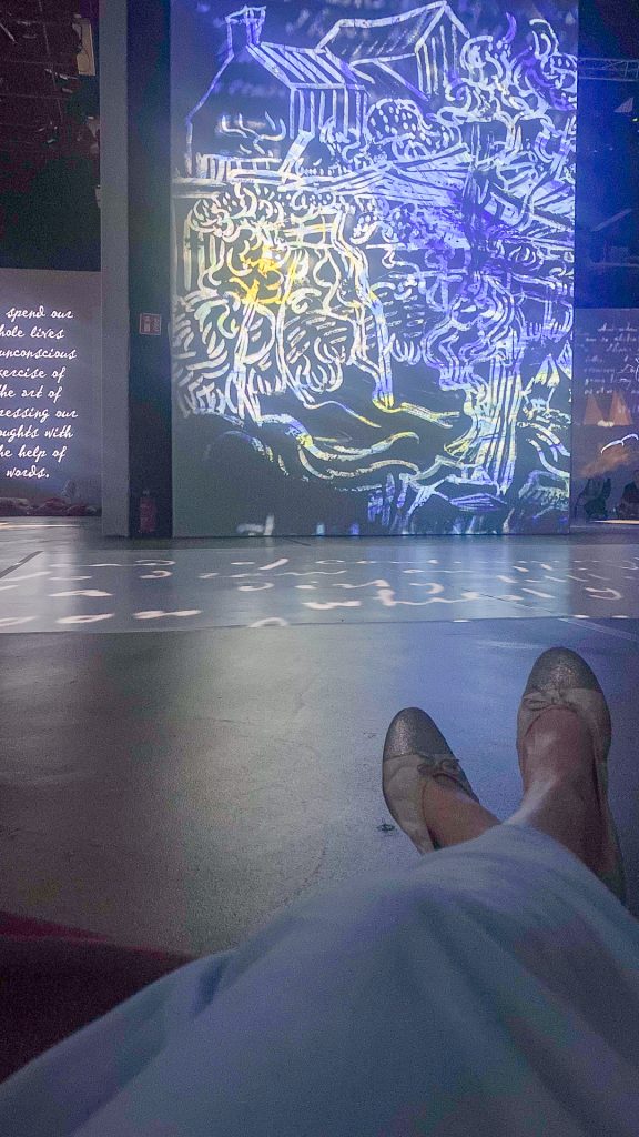 Lähikuva henkilön jaloista ja kengistä, taustalla heijastuu Van Goghin taidenäyttelyn teoksia. Kuva symboloi taiteellista inspiraatiota lomalla.