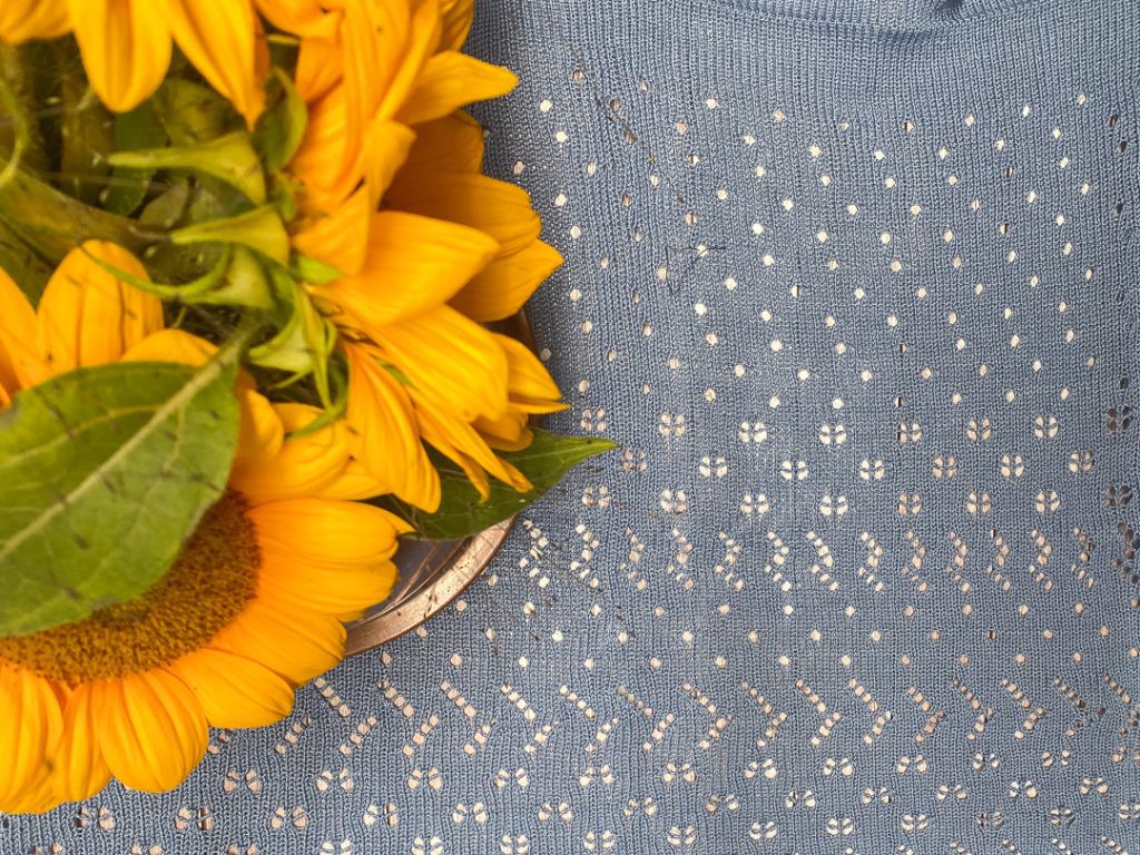 Lähikuva kirkkaankeltaisista auringonkukista, jotka lepäävät vaaleansinisellä, pitsineuloksella koristellulla neuloksella Kuva symboloi luonnon ja käsityön liittoa.