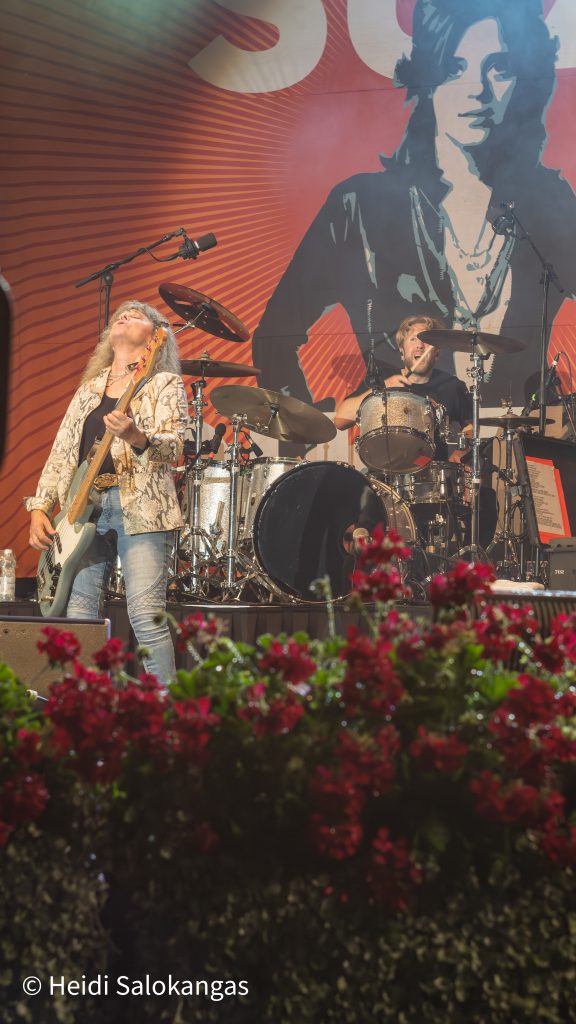 Suzi Quatro esiintyy lavalla soittaen bassoa, taustalla hänen bändikaverinsa.