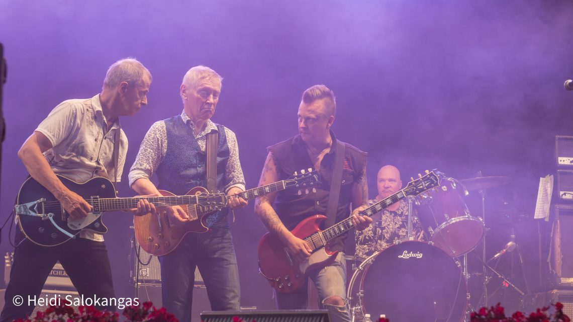 Kolme kitaristia soittaa lavaesiintymisessä, edessä Black Devils -yhtyeen kitaristi, vierellään Hurriganesin alkuperäiset kitaristit. Kuva on otettu matalalta yleisön tasolta.