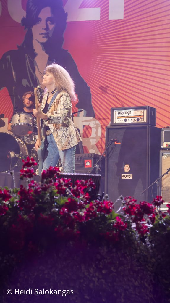 Suzi Quatro esiintyy lavalla soittaen bassoa, taustalla hänen kuvansa.