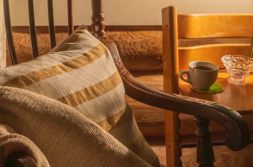 Lämminhenkinen lähikuva lukunurkkauksesta, jossa näkyy perinteinen puinen tuoli, lämmin peitto ja raidallinen tyyny. Taustalla kahvikuppi lasinalusen päällä.