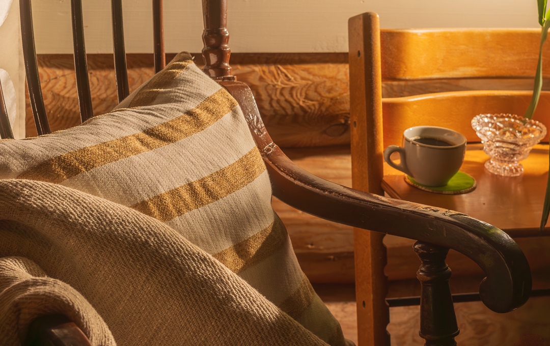 Lämminhenkinen lähikuva lukunurkkauksesta, jossa näkyy perinteinen puinen tuoli, lämmin peitto ja raidallinen tyyny. Taustalla kahvikuppi lasinalusen päällä.