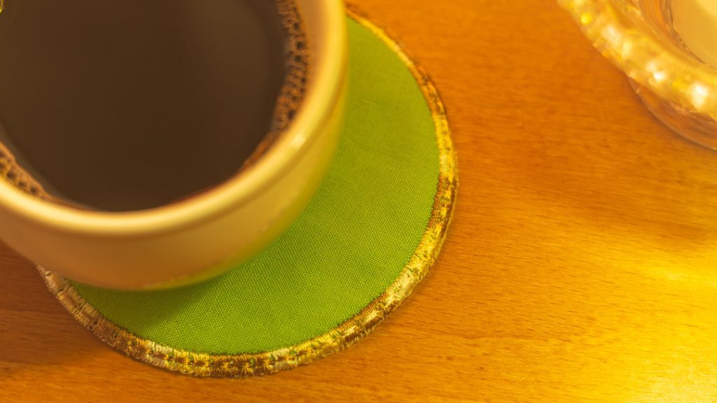 Lähikuva kahvikupista, joka seisoo pyöreän, kirkkaan vihreän lasinalusen päällä puupöydällä. Lasinalusen reunat on kirjailtu kullanvärisellä langalla.