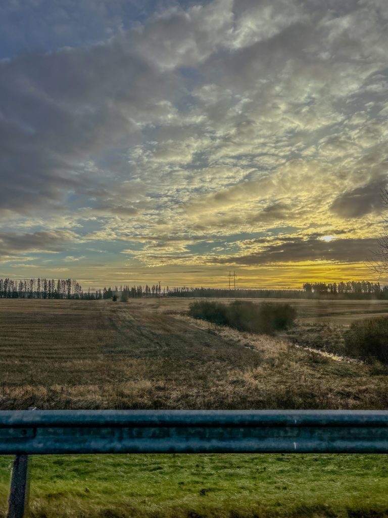 Kuva on otettu auton ikkunasta, ja siinä näkyy laaja pelto sekä matalalta paistavan auringon pehmeä valo. Taivaan pilvet hehkuvat vaaleankeltaisina ja sinertävinä. Maisema muistuttaa niistä rauhallisista väleistä, joita matkanteko tarjoaa kiireisen arjen keskellä.