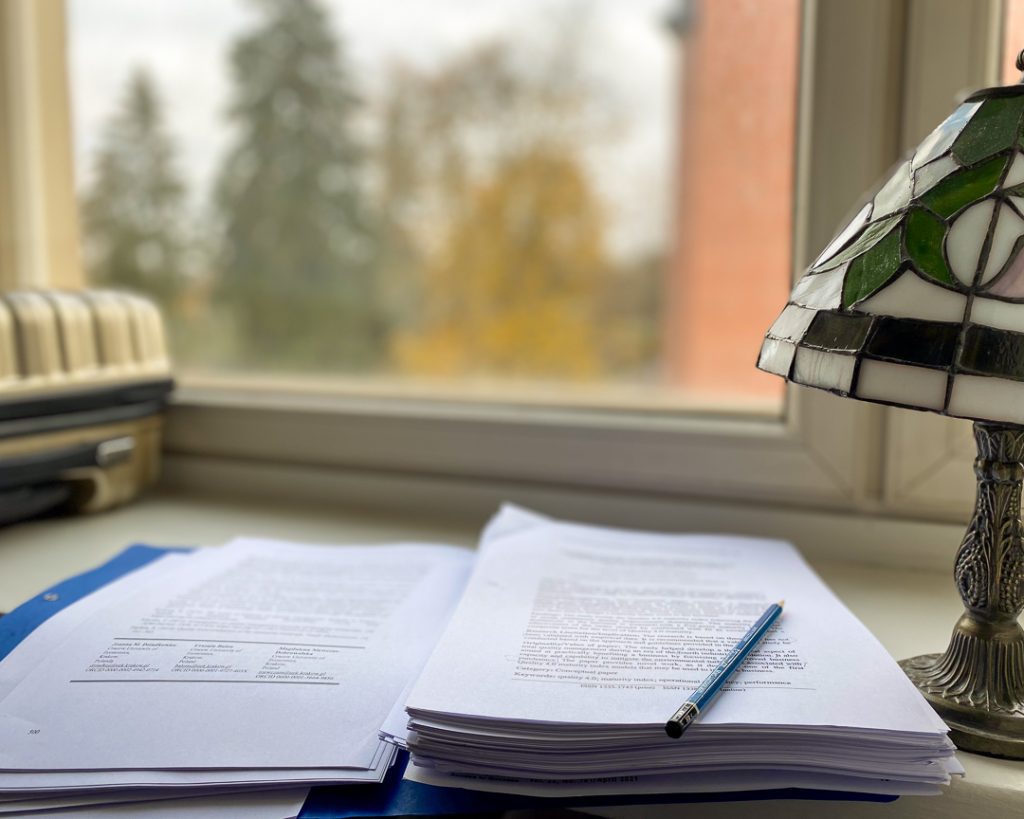Tutkimusartikkeleita ja lyijykynä ikkunalaudalla, taustalla syksyinen maisema – marraskuinen työhetki ilman tietokonetta.
