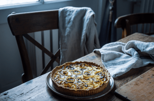Gluteeniton ja vegetaarinen Quiche Lorraine yksinkertaisella keraamisella lautasella puupöydällä. Luonnonvalo ja pohjoismainen keittiö luovat rauhallisen, nostalgisen tunnelman.