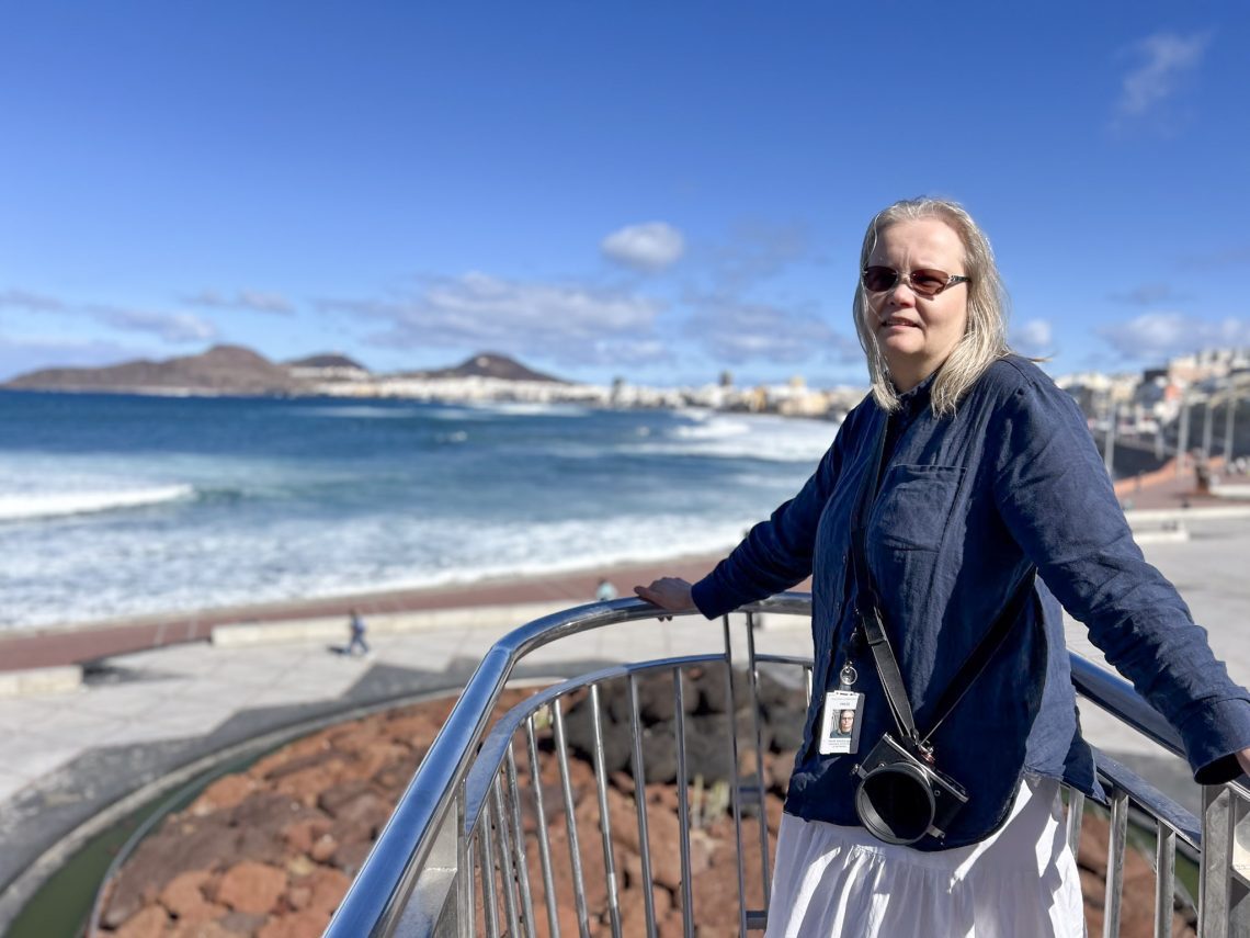Nainen seisoo kaiteella Atlantin rannalla Las Palmasissa, kamera kaulassa ja aurinkolasit silmillä. Taustalla näkyy kaupungin rantaviiva ja vuoria. Kuvassa käsityö- ja kirjoittajavaikuttaja keräämässä inspiraatiota