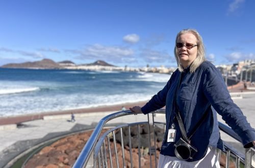 Nainen seisoo kaiteella Atlantin rannalla Las Palmasissa, kamera kaulassa ja aurinkolasit silmillä. Taustalla näkyy kaupungin rantaviiva ja vuoria. Kuvassa käsityö- ja kirjoittajavaikuttaja keräämässä inspiraatiota