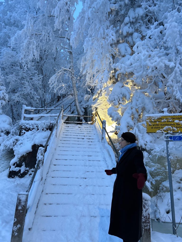 umipeitteiset portaat nousevat talviseen metsään; tummaan takkiin pukeutunut henkilö punaisine rukkasineen seisoo portaiden juurella, ympärillä paksut lumiset oksat ja lämmin valonhehku.