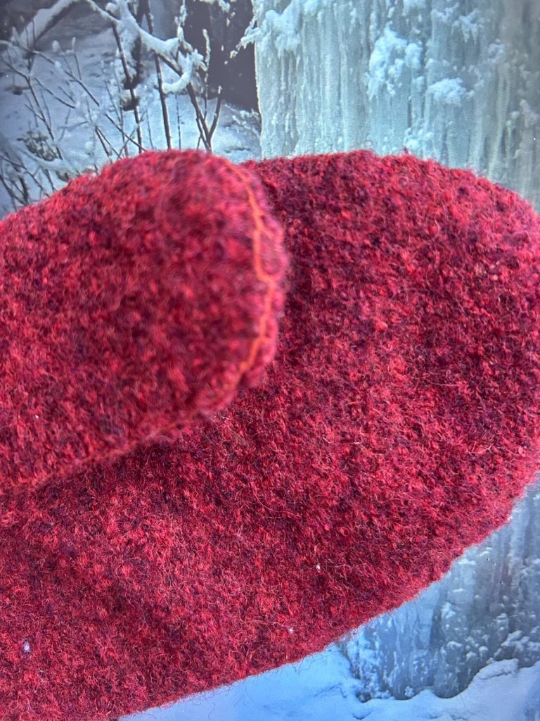 Kuvassa näkyy lähikuvassa punaisesta villaneuloksesta ommellut naisten lapaset. Materiaali on tiivistä ja huopamaista, ja lapasen saumalinja kulkee selkeästi sivua pitkin. Taustalla on talvinen maisema, jossa näkyy jäätä ja lunta, mikä korostaa lapasten käyttötarkoitusta kylmissä olosuhteissa. Kuva välittää käsityön lämpöä, käytännöllisyyttä ja talvista rauhaa.