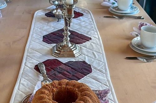 Katettu pöytä, jonka keskellä kulkee valkoinen tikattu kaitaliina sydänapplikaatioilla. Hopeinen kynttilänjalka ja kukkakimppu luovat juhlatunnelman, ja kahvikakku viimeistelee asetelman.