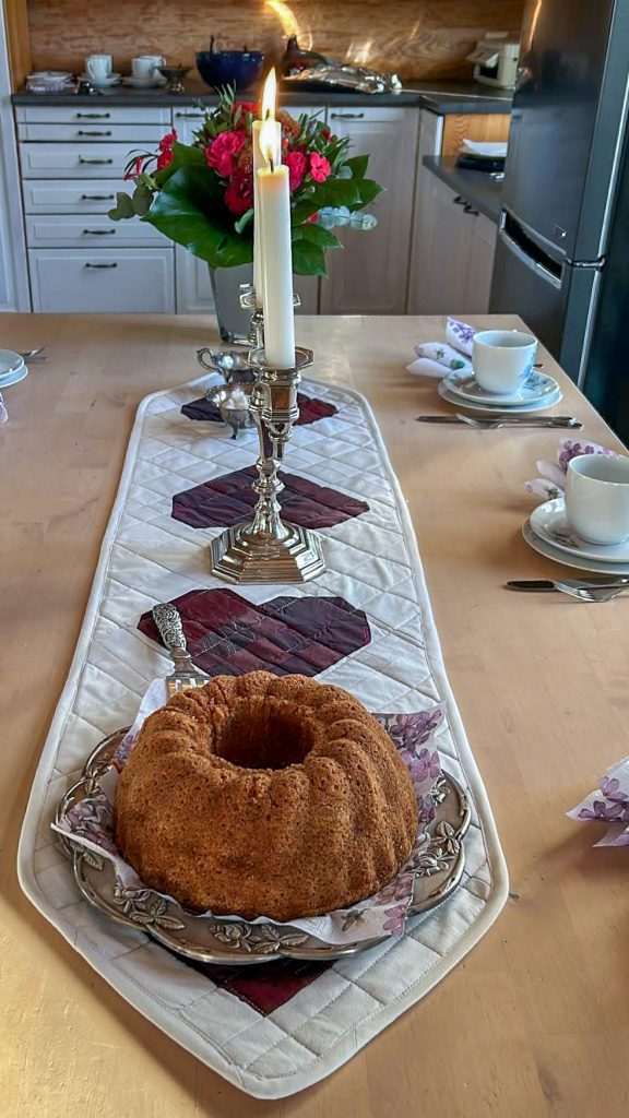 Katettu pöytä, jonka keskellä kulkee valkoinen tikattu kaitaliina sydänapplikaatioilla. Hopeinen kynttilänjalka ja kukkakimppu luovat juhlatunnelman, ja kahvikakku viimeistelee asetelman.