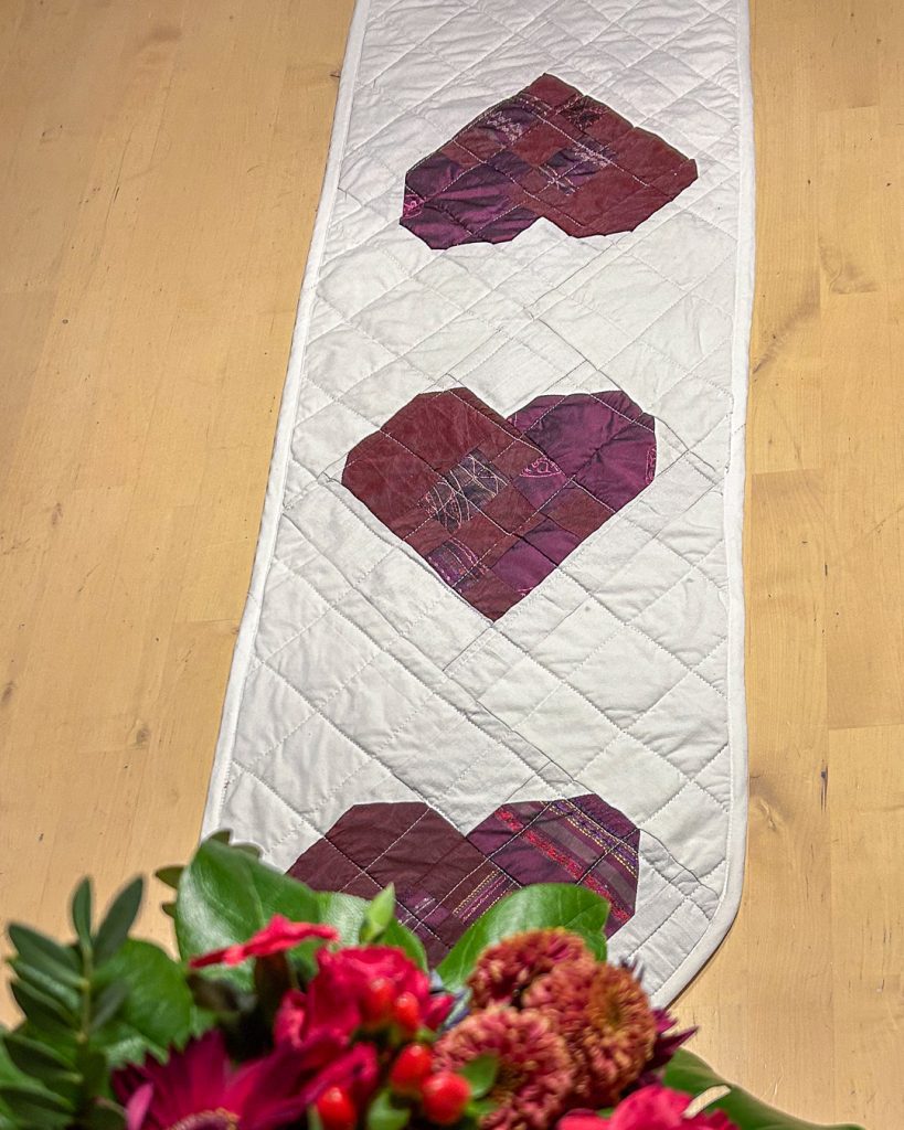 Sydänaiheinen tilkkutyö kaitaliina valkoisella taustalla, kolme sydäntä kierrätyskankaista ommeltuna.