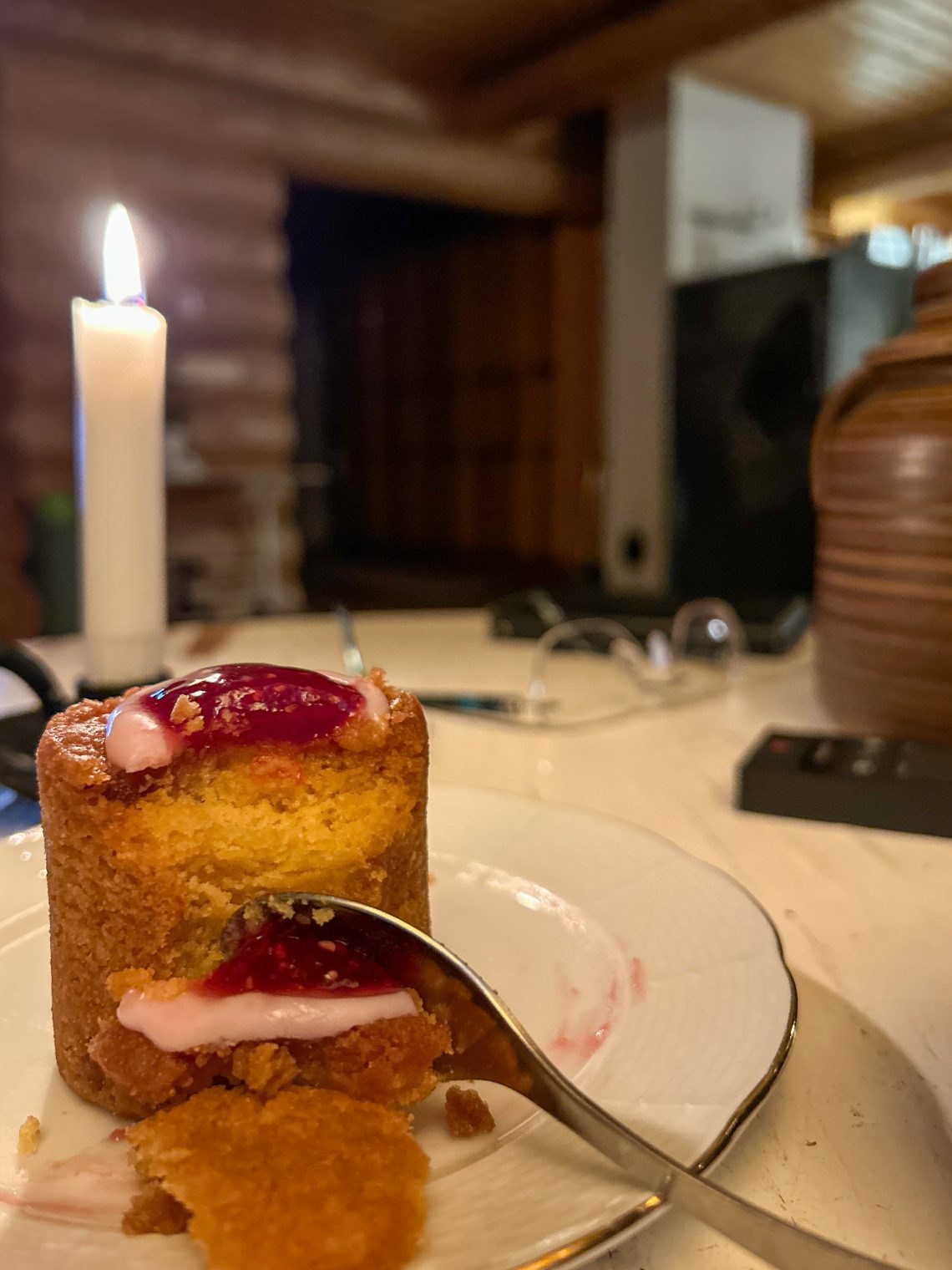 Runebergin torttu jamilla ja vaaleanpunaisella kuorrutteella valkoisella lautasella; vieressä ruskea teepannu ja palava kynttilä rustiikkisessa ympäristössä.