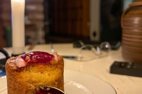 Runebergin torttu jamilla ja vaaleanpunaisella kuorrutteella valkoisella lautasella; vieressä ruskea teepannu ja palava kynttilä rustiikkisessa ympäristössä.