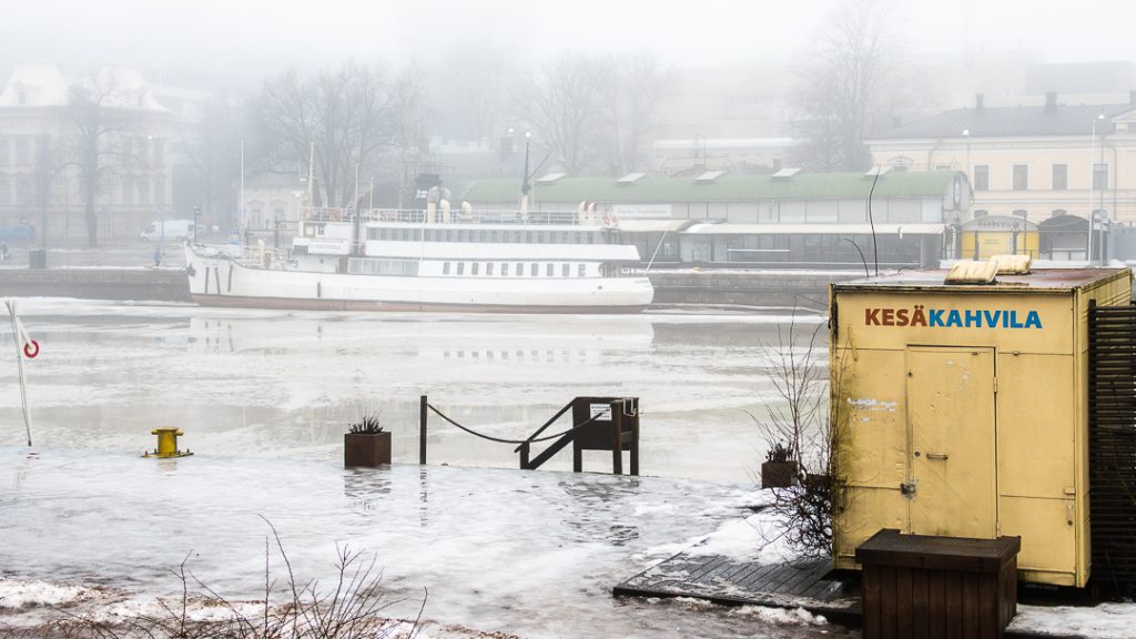 Sumuinen Aurajoki Turussa: jokiranta, vanha laiva ja keltainen kesäkahvilarakennus harmaassa talvisäässä.