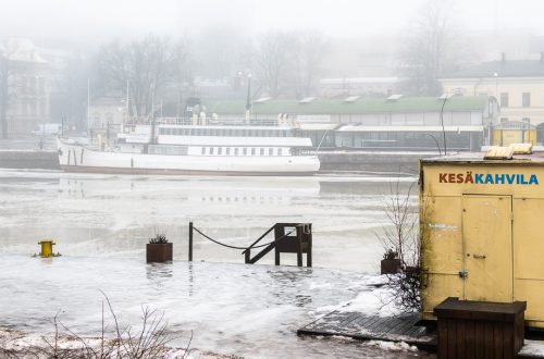 Sumuinen Aurajoki Turussa: jokiranta, vanha laiva ja keltainen kesäkahvilarakennus harmaassa talvisäässä.