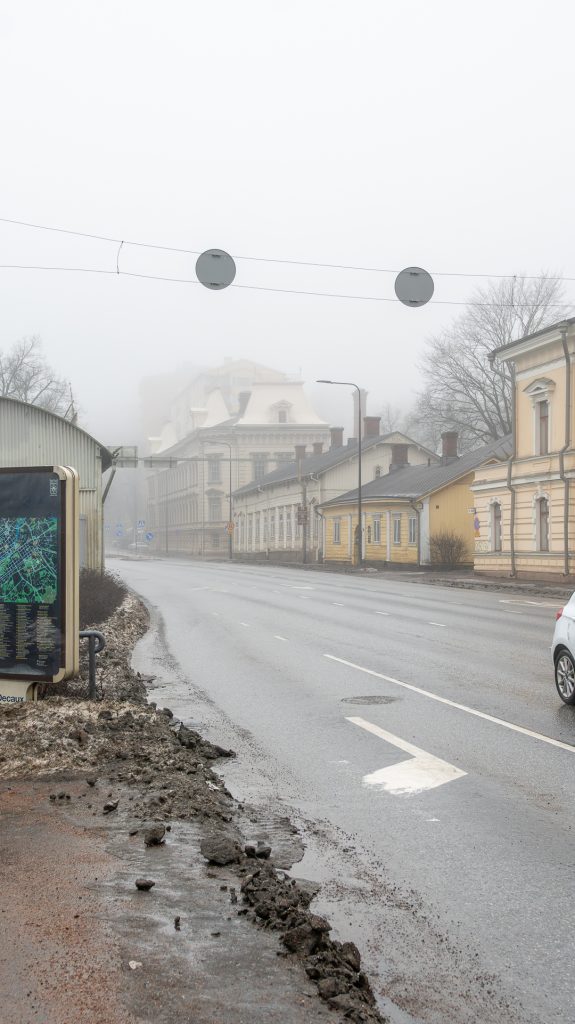 Sumuinen katunäkymä Turussa: keltainen puutalo, hiljainen tie ja pysäkkikartta aamuhämärässä.