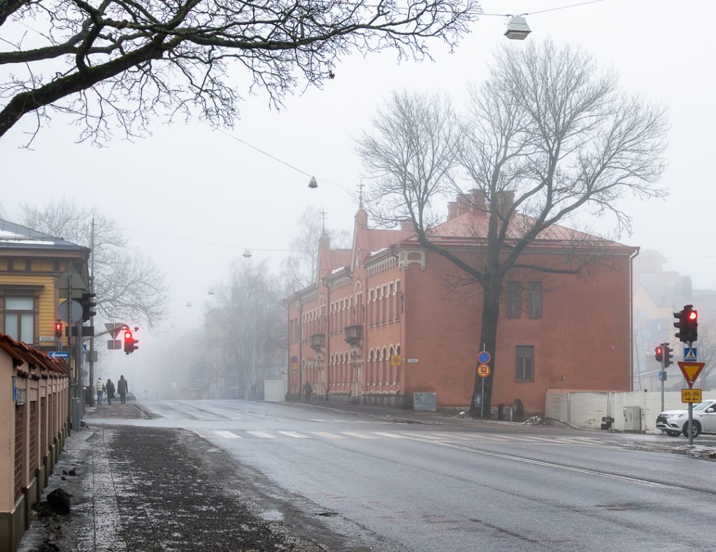 Sumuinen katunäkymä Turussa: punaruskea historiallinen rakennus, iso lehdettömäksi jäänyt puu ja punaiset liikennevalot hiljaisella kadulla.