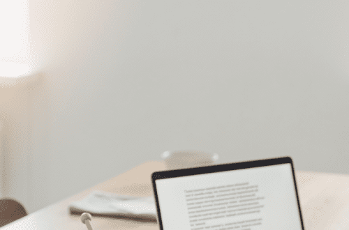 A calm Nordic editorial hero image showing a wooden table with yarn, knitting needles, fabric pieces and a laptop with a text document open, soft natural window light, minimal composition, lots of negative space, thoughtful atmosphere, magazine feature photography, muted natural colors