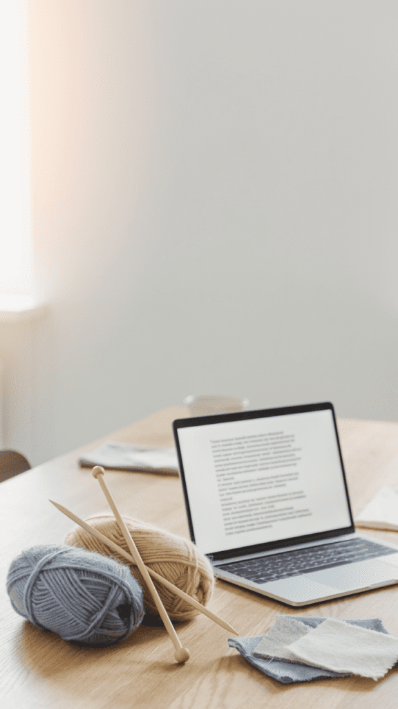 A calm Nordic editorial hero image showing a wooden table with yarn, knitting needles, fabric pieces and a laptop with a text document open, soft natural window light, minimal composition, lots of negative space, thoughtful atmosphere, magazine feature photography, muted natural colors